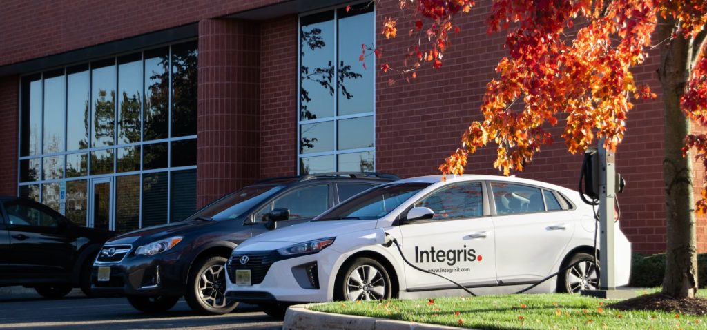 The exterior of our Cranbury office. A brick building with large windows with the Integris logo above. An electric car with the Integris logo on the side is charging. To the right, a tree is bright red in autumn.