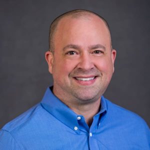 Professional headshot of a smiling man wearing a blue shirt against a gray background.