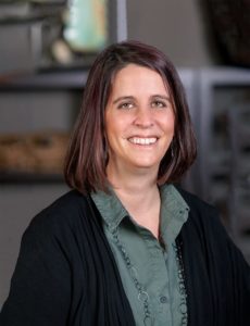 A portrait of a smiling woman with shoulder-length brown hair, wearing a black cardigan, green blouse, and a necklace.