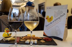Two wine glasses with red and white wine next to a platter of cheese, crackers, and dried meats. A flyer with a duck image in the background reads Don't Be A Sitting Duck.