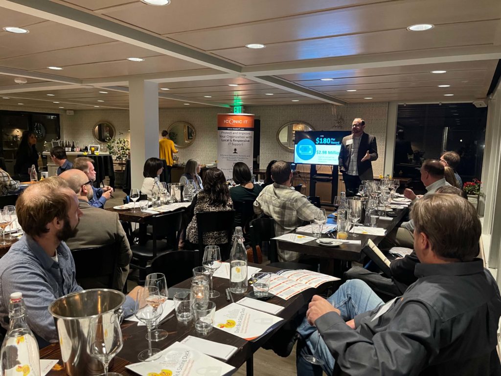 A man gives a presentation to a group of people seated at tables in a well-lit restaurant. A projector screen displays a slide, and attendees have papers and glasses in front of them.