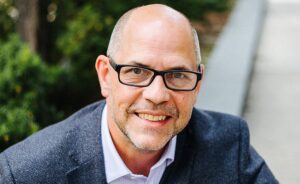 A man with glasses and a beard smiles while wearing a dark blazer and light-colored shirt outdoors.
