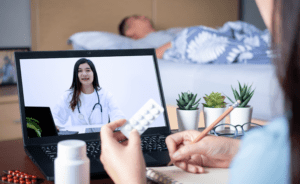 A person holds a blister pack and writes notes while having a telemedicine consultation with a doctor on a laptop. A bed and various plants are in the background.