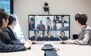 A group of people in a conference room participate in a video call with colleagues displayed on a large screen in front of them.
