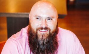 A bearded man smiles at the camera with blurred background.