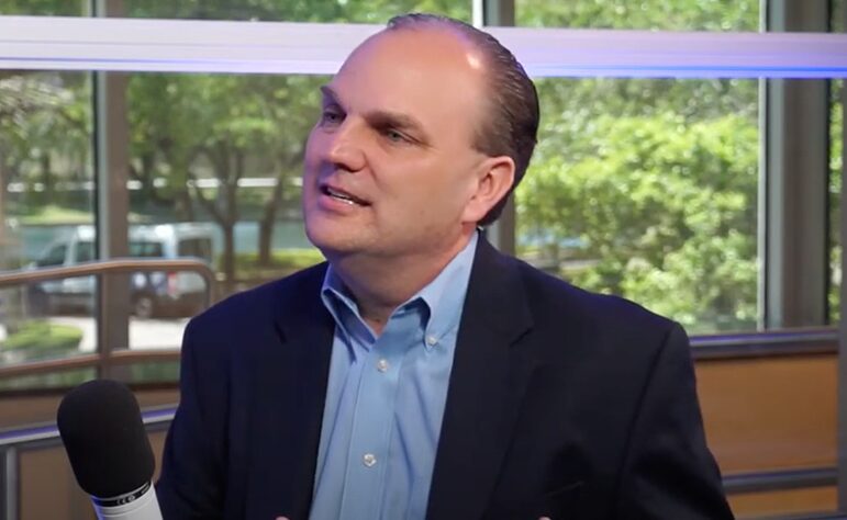 A man in a dark blazer and light blue shirt addresses an interviewer while holding a microphone. The background shows a window with a green outdoor view.