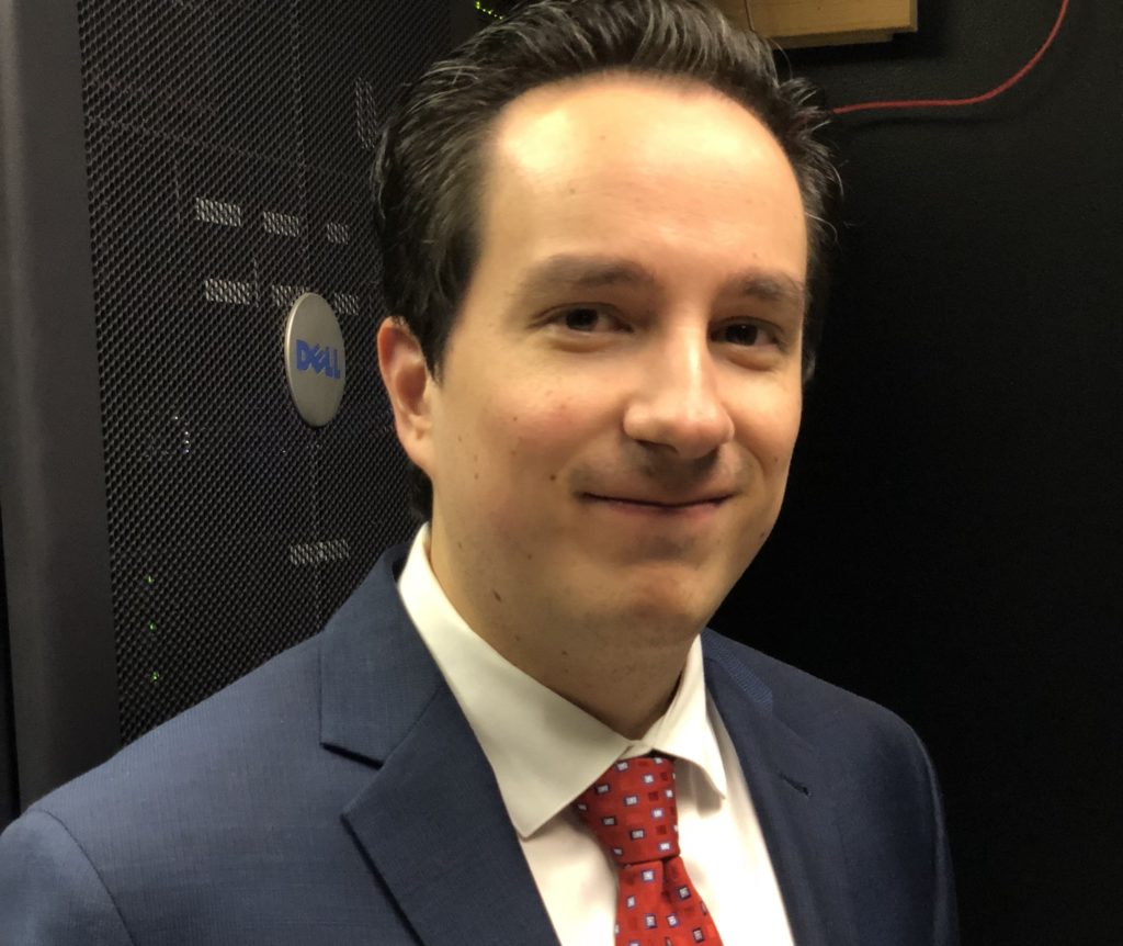 A person in a blue suit and red tie stands in front of an Iconic IT Dell server rack.