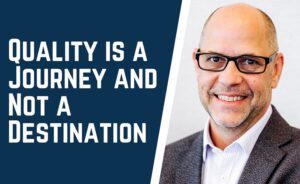 Man smiling with glasses in a suit on the right, text on the left reads QUALITY IS A JOURNEY AND NOT A DESTINATION.