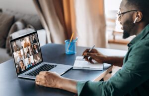 A person wearing earphones sits at a desk, diligently taking notes while attending a video conference on a laptop. Multiple participants are visible on the laptop screen.