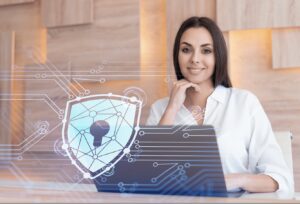 A woman is sitting at a desk with a laptop, her hand resting on her chin. A digital shield with a keyhole and network lines is superimposed on the image, symbolizing cybersecurity.