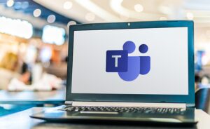 A laptop on a table with the Microsoft Teams logo displayed on the screen. The background is blurred, featuring people and lights.