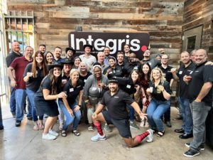 A large group of people posing together in front of a wooden wall with a sign that reads Integris. Many are holding drinks and smiling, celebrating employee recognition.