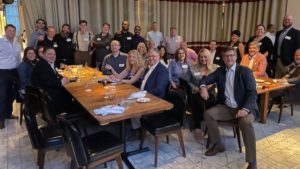 A group of people from an IT solution provider pose together around two wooden tables in a restaurant setting. Many are seated while others stand behind, smiling and wearing name tags.