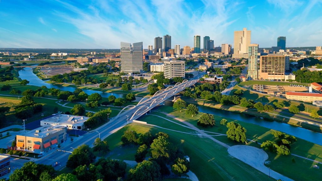 Aerial view of Fort Worth featuring high-rise buildings, a prominent bridge, and green spaces with trees, under a blue sky with scattered clouds.