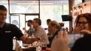 A group of people sitting at a wooden bar. A woman with curly hair and glasses laughs while a man in an Integris shirt serves a drink, proudly showcasing his affiliation with the IT solution provider. Some patrons in the background are engaged in conversation.