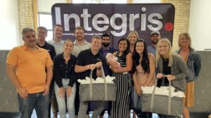 A group of people standing in front of an Integris banner, an IT solution provider. Two individuals in the center hold bags, and one person holds a baby. They are all smiling.