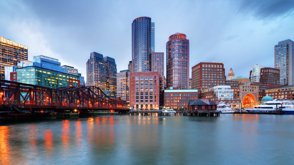 Panoramic view of Boston's waterfront at dusk, featuring illuminated buildings, a red bridge, and boats docked along the harbor.