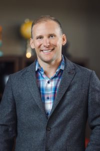 A smiling man in a grey blazer and checkered shirt posing for a portrait, known as John Marinac.