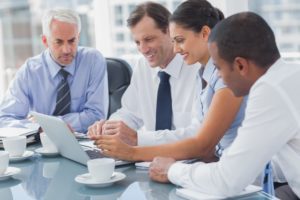 Four business professionals in formal attire are seated around a table, engaged in a discussion while looking at a laptop screen.