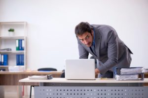 A man in a gray suit leans over a desk, looking at a laptop. On the desk are tall stacks of papers. The background features shelves with folders and office supplies.