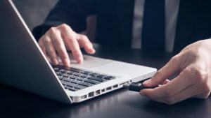 A person inserts a USB flash drive into a laptop on a dark table, with one hand on the keyboard and the other holding the flash drive.