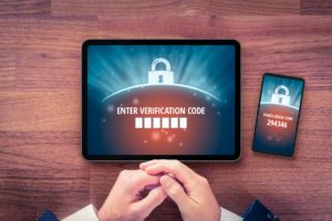 A tablet and smartphone display a verification code entry screen. Both devices show a lock icon and verification code numbers. Hands are seen resting on a wooden table.