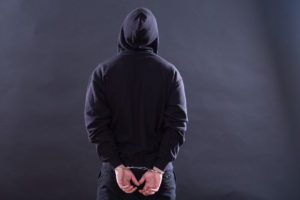A person wearing a black hoodie, seen from behind, stands with their hands cuffed behind their back against a dark background.
