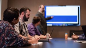 Group of IT Services professionals engaged in a presentation with a speaker pointing at a screen.
