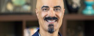 A man with a mustache and goatee wearing a blue suit jacket smiles confidently at the camera, ready for press coverage.