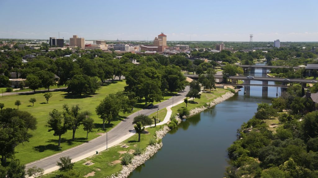 Aerial view of a river flowing through a city with numerous buildings, large grassy areas, and abundant trees on a sunny day.