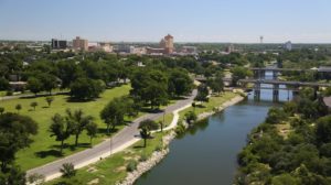 Aerial view of a river flowing through a city with numerous buildings, large grassy areas, and abundant trees on a sunny day.