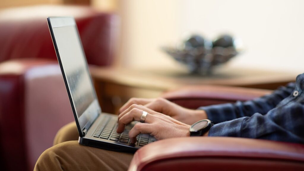 Person using a laptop while sitting in a red chair, with a bowl of decorative items in the background.