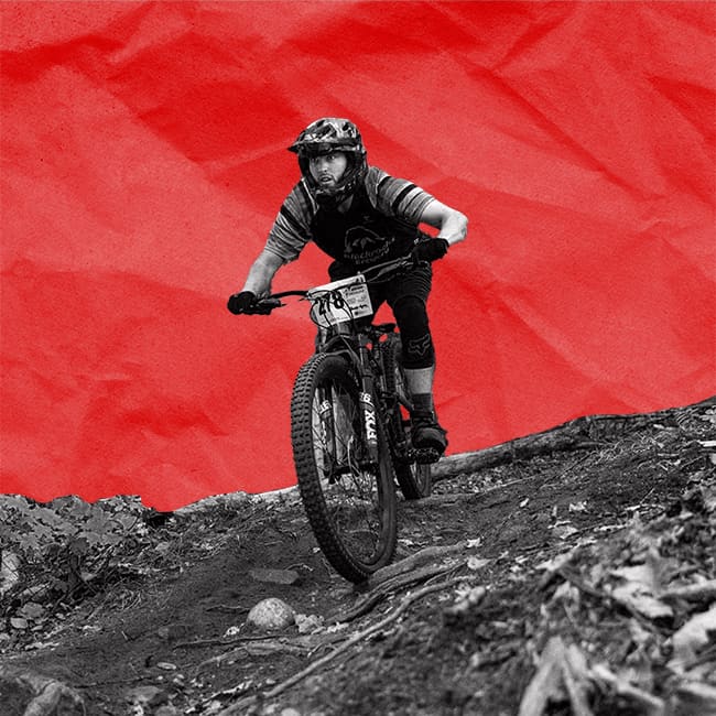 A mountain biker rides downhill on a rugged trail, wearing a helmet and protective gear. The background is a textured red.