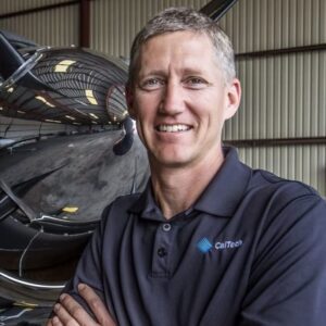 A man wearing a dark polo shirt with a "CalTech" logo stands in front of an aircraft inside a hangar, arms crossed and smiling.