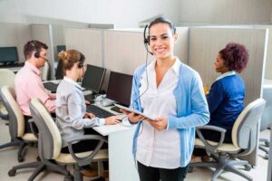 A smiling woman wearing a headset and holding a tablet stands in the foreground of an office with other employees working at computer stations.