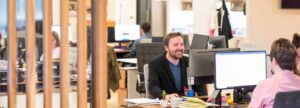 People working at desks with computer monitors in an open-plan office; one man is smiling and conversing with a colleague.