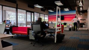 A modern office space with people working at desks, a man writing on a wall, and colorful furniture. Large windows provide natural light.