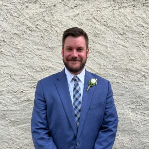A man wearing a blue suit, white shirt, and checkered tie smiles while standing in front of a textured light-colored wall. He has a boutonniere pinned to his left lapel.