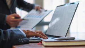 Close-up of a person's hand typing on a laptop keyboard with another person holding a clipboard with charts in the background. A notebook and pen are on the desk.