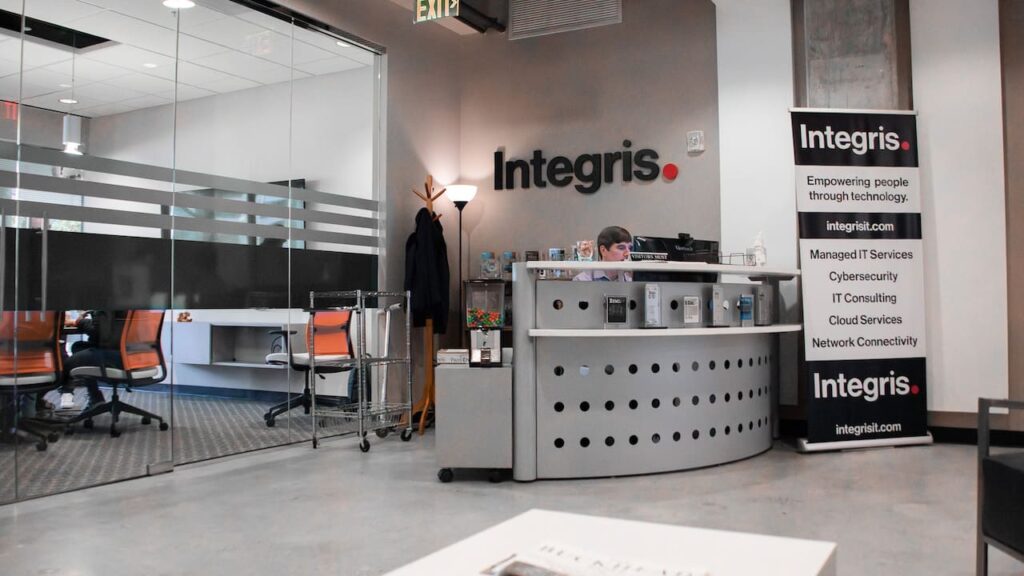 A modern office reception area for Integris features a receptionist at a sleek, curved desk with branding signage. The background includes glass-walled meeting rooms with orange chairs. The atmosphere is clean and professional.