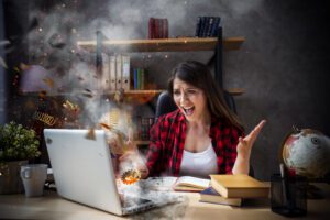A woman wearing a red plaid shirt sits at a desk, looking frustrated as her laptop appears to be overheating, with springs and gears flying out. Her arms are raised in exasperation. The desk has books, a globe, a plant, and other office supplies on it.
