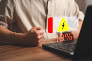 A person sits at a desk with a laptop. A low battery warning symbol, showing a lightning bolt and "10%" displayed in red, hovers above the screen. The individual's hand is clenched into a fist, possibly revealing frustration as the laptop battery drains rapidly.