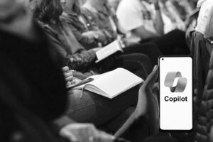 Black and white photo showing an audience seated close together, taking notes. In the foreground, a person's hand holding a smartphone with the "Copilot" logo displayed on the screen, hinting at seamless it support in action.