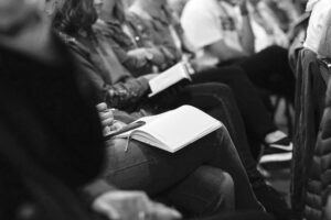 Black and white photo of people sitting in a row, possibly at a meeting or lecture. The focus is on their hands, some holding pens and notebooks, while others rest on their laps. The attendees, likely part of an IT support team, are dressed casually, suggesting an informal or relaxed environment.