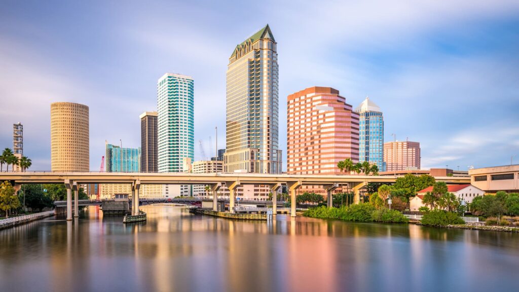 A downtown cityscape featuring several skyscrapers of varying heights and designs, including a prominent triangular-roofed building. Below them is a calm river with reflections of the buildings, crossed by an elevated road with lush greenery on the riverbanks.