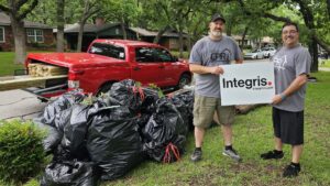Two men wearing grey t-shirts stand on a lawn next to a red pickup truck loaded with wooden planks. They are holding a large sign that reads "Integris" with a website link. Piles of black garbage bags are on the ground in front of them, indicating a cleanup effort.