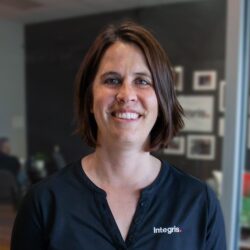 A woman with short brown hair, wearing a black shirt with the "Integris" logo, stands indoors and smiles at the camera.