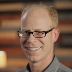 A man with light skin, short blond hair, and black glasses smiles at the camera. He is wearing a gray shirt, with a blurred background of horizontal wooden panels.