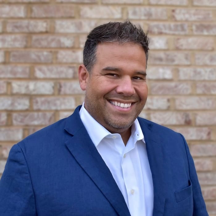 A man with short dark hair and a beard, exuding leadership in a blue suit and white shirt, smiles confidently in front of a brick wall.