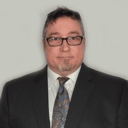 A man with short dark hair and glasses, wearing a dark suit, white shirt, and patterned tie, poses against a plain light background.